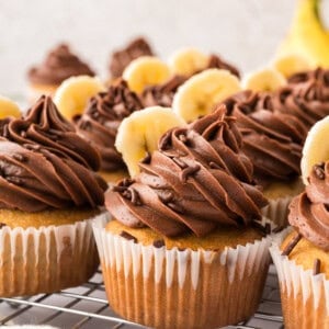 Banana cupcakes with chocolate frosting and banana slices on top, arranged on a cooling rack.