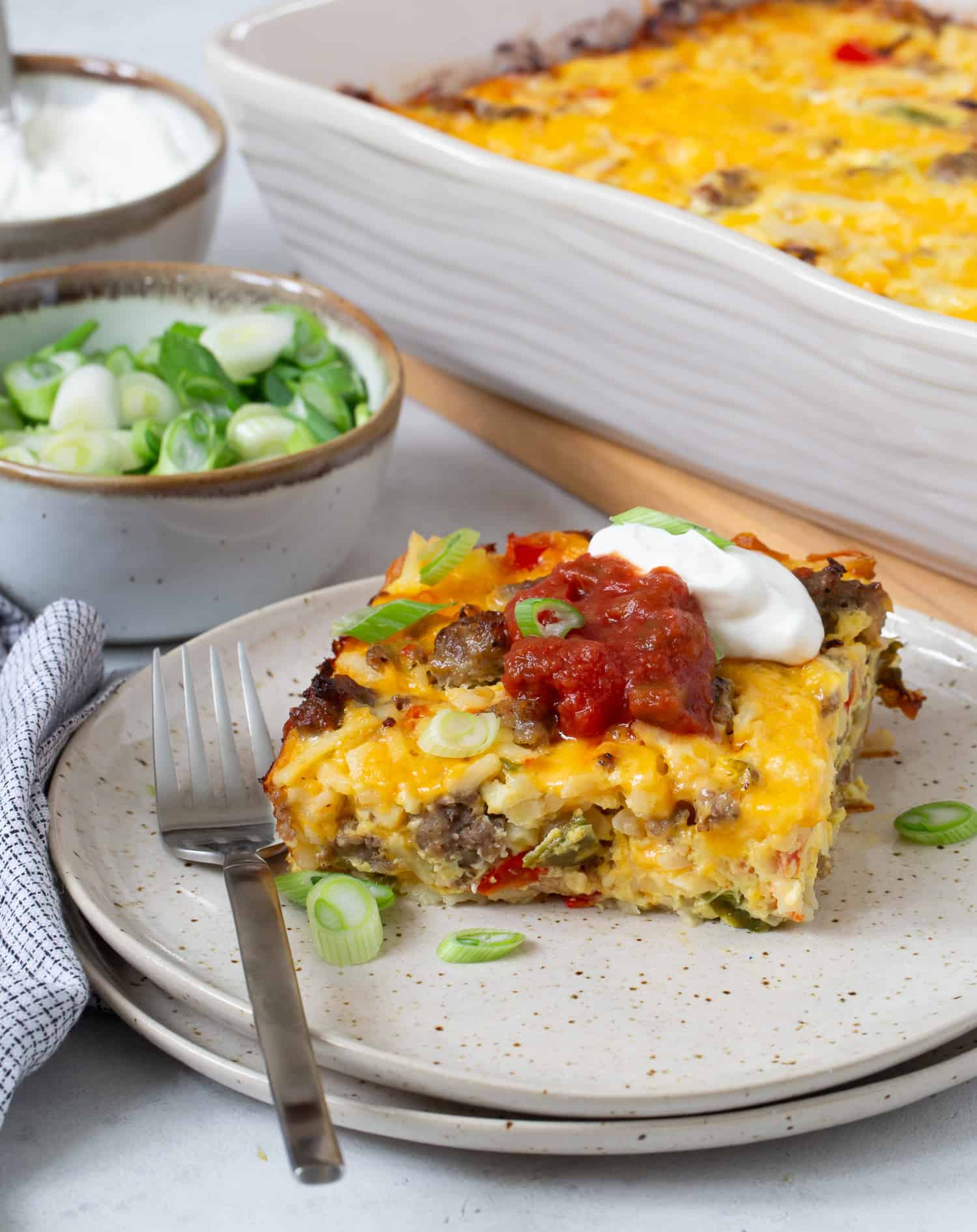 Slice of hash brown breakfast casserole topped with salsa and sour cream, remaining casserole in background.