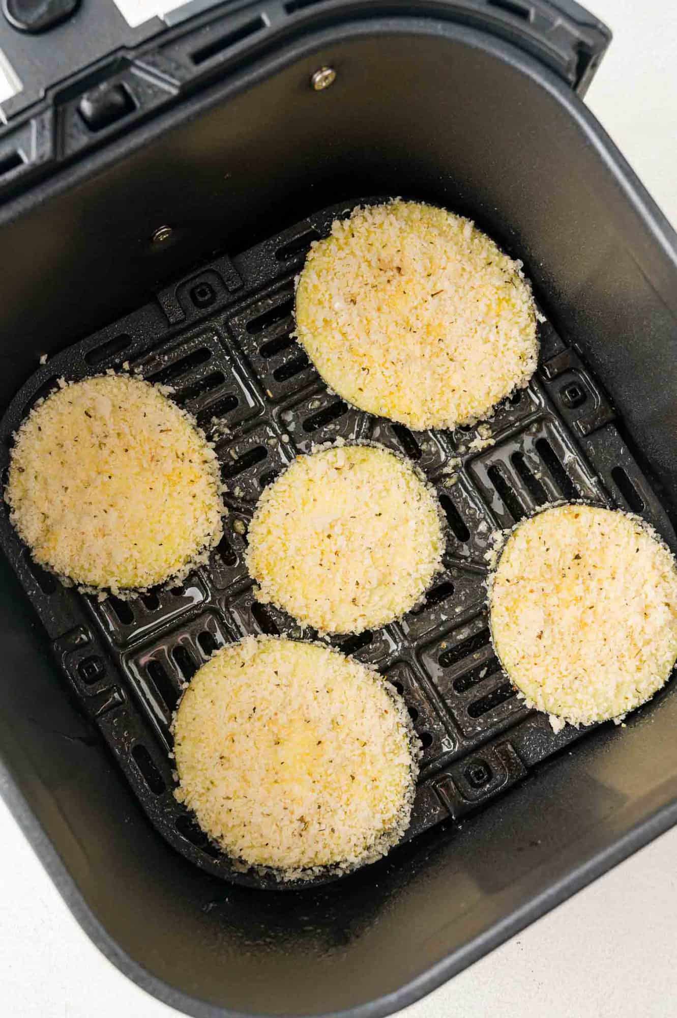 Breaded, uncooked eggplant rounds in air fryer basket.