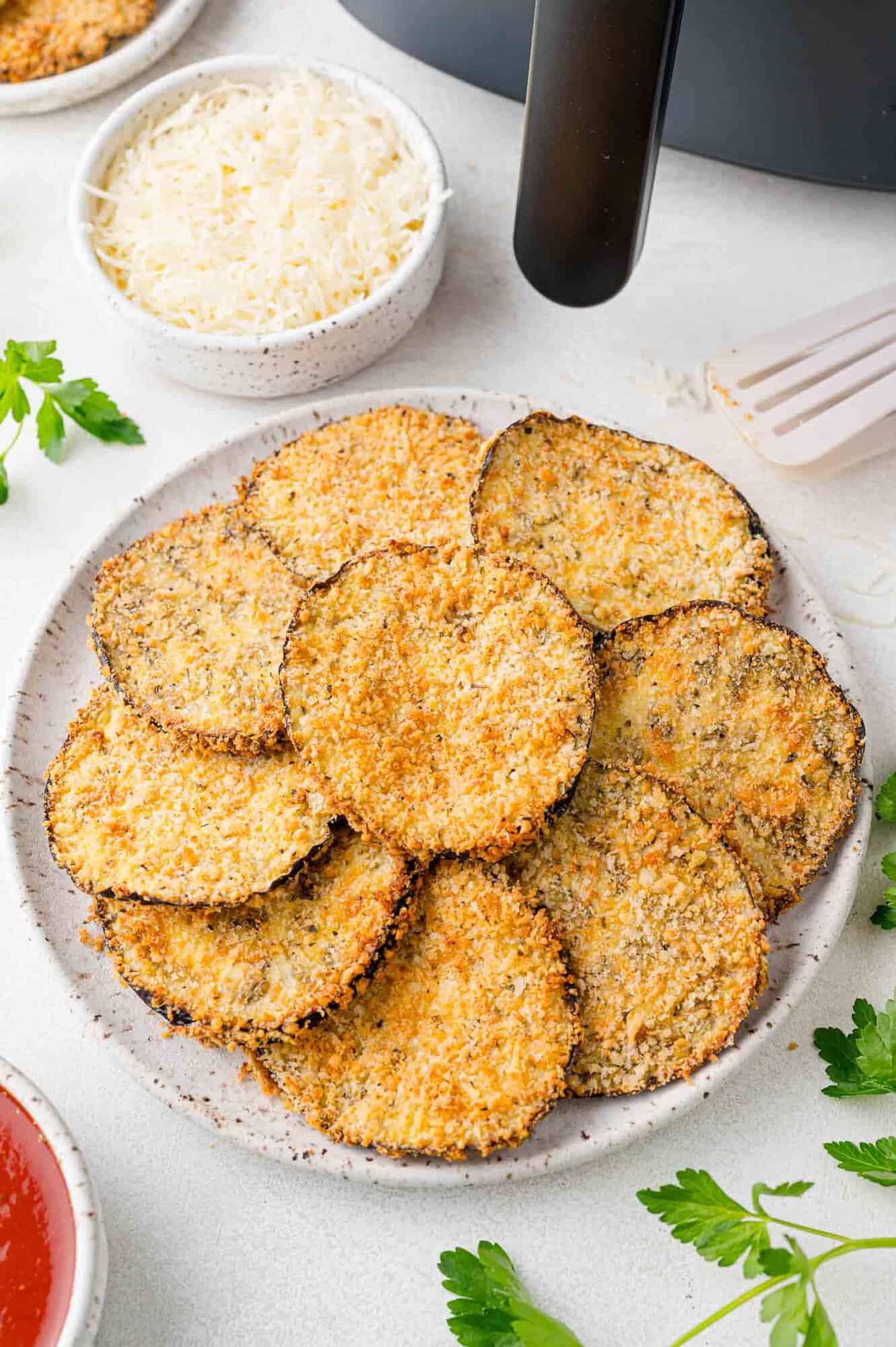 Crispy eggplant slices arranged on a plate, air fryer also present in corner of photo.