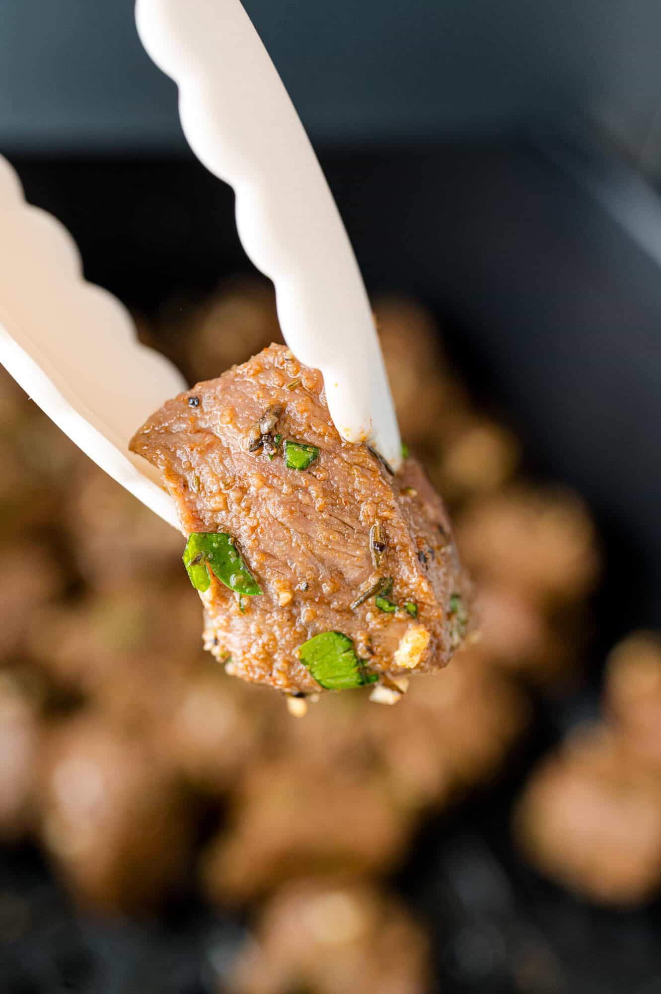 Air fryer steak bite held with tongs over more steak bites.