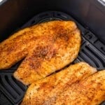 Two seasoned pieces of air fryer tilapia still in air fryer basket.