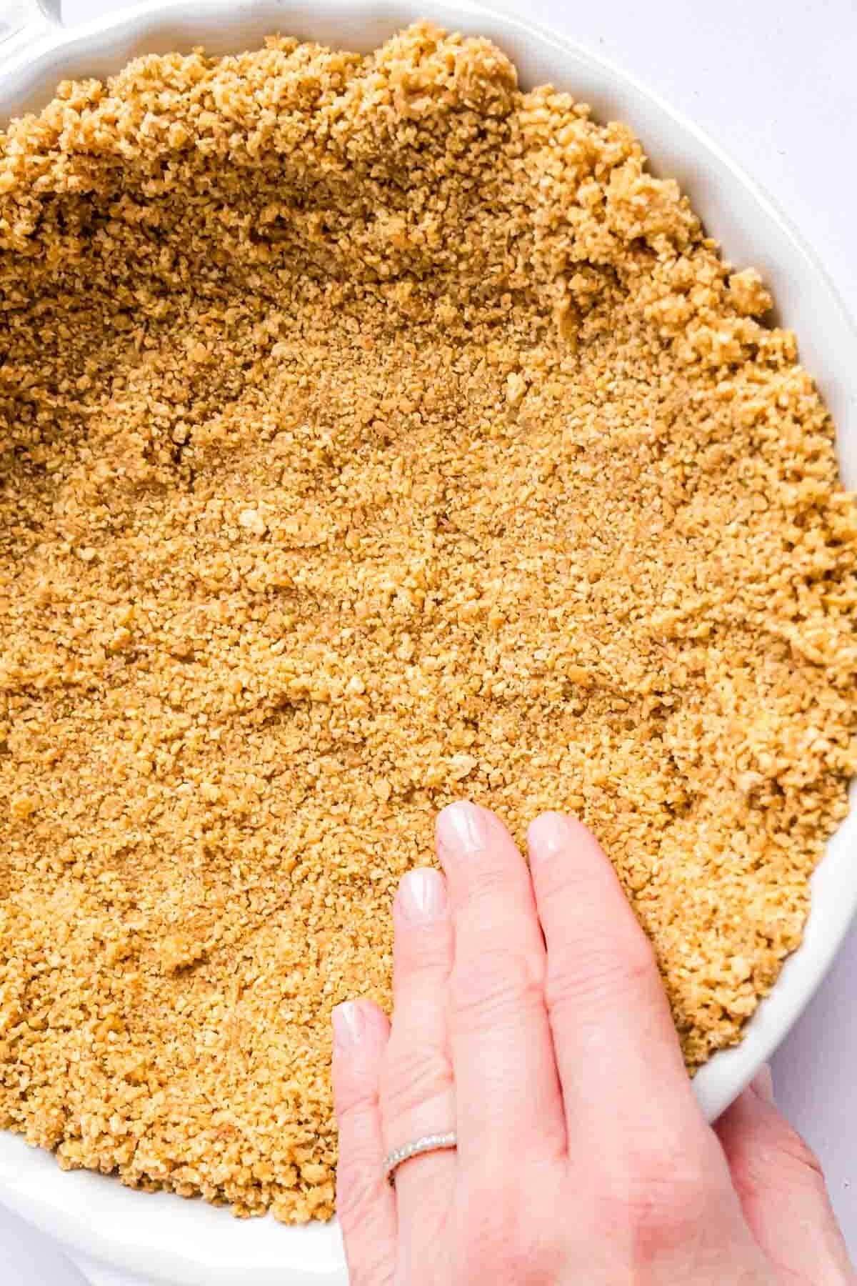 A hand pressing graham cracker crumb mixture into a pie plate.