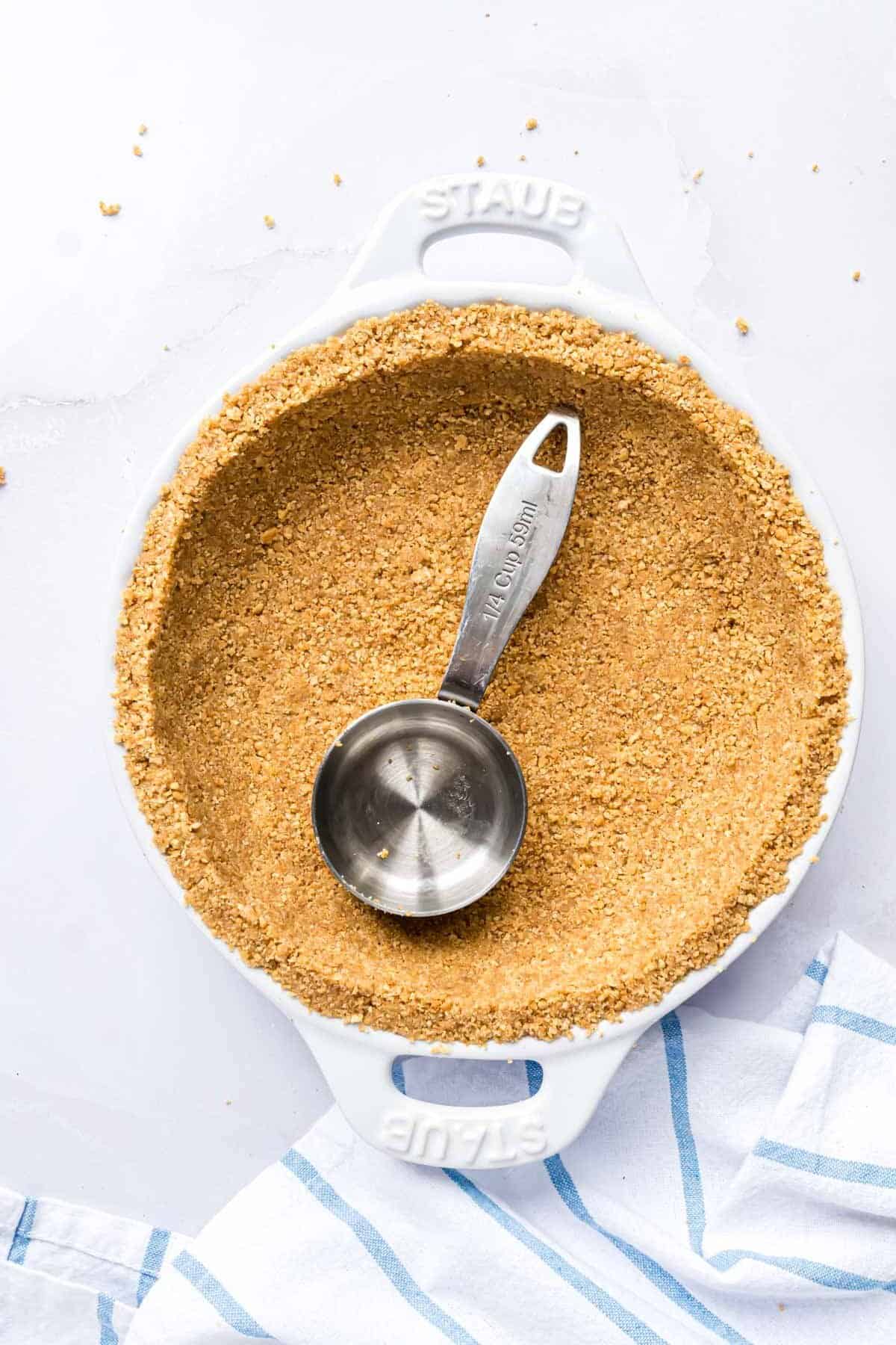 A measuring cup being used to press graham cracker crust into a pie plate.