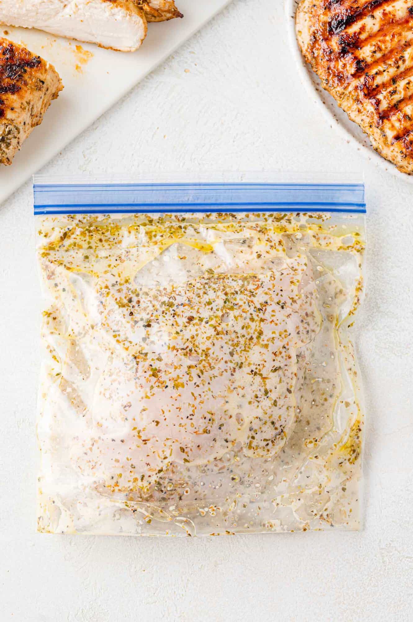 Chicken in Greek chicken marinade, in a zip-top bag, surrounded by grilled chicken.