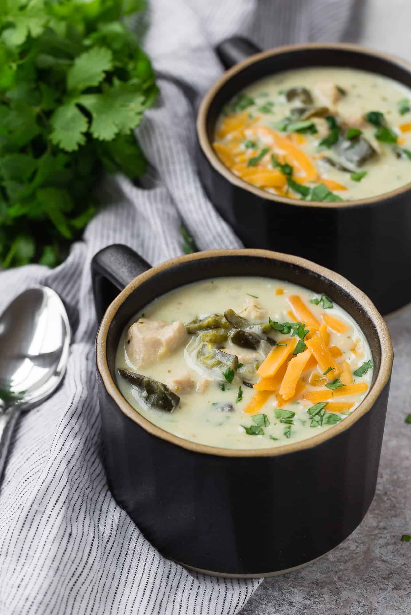 Two mug-style bowls of chicken chile relleno soup, topped with cheddar.