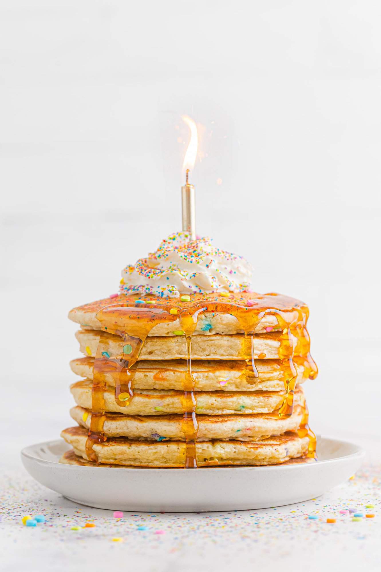 Stack of birthday cake pancakes, with syrup, with a lit birthday candle on top.