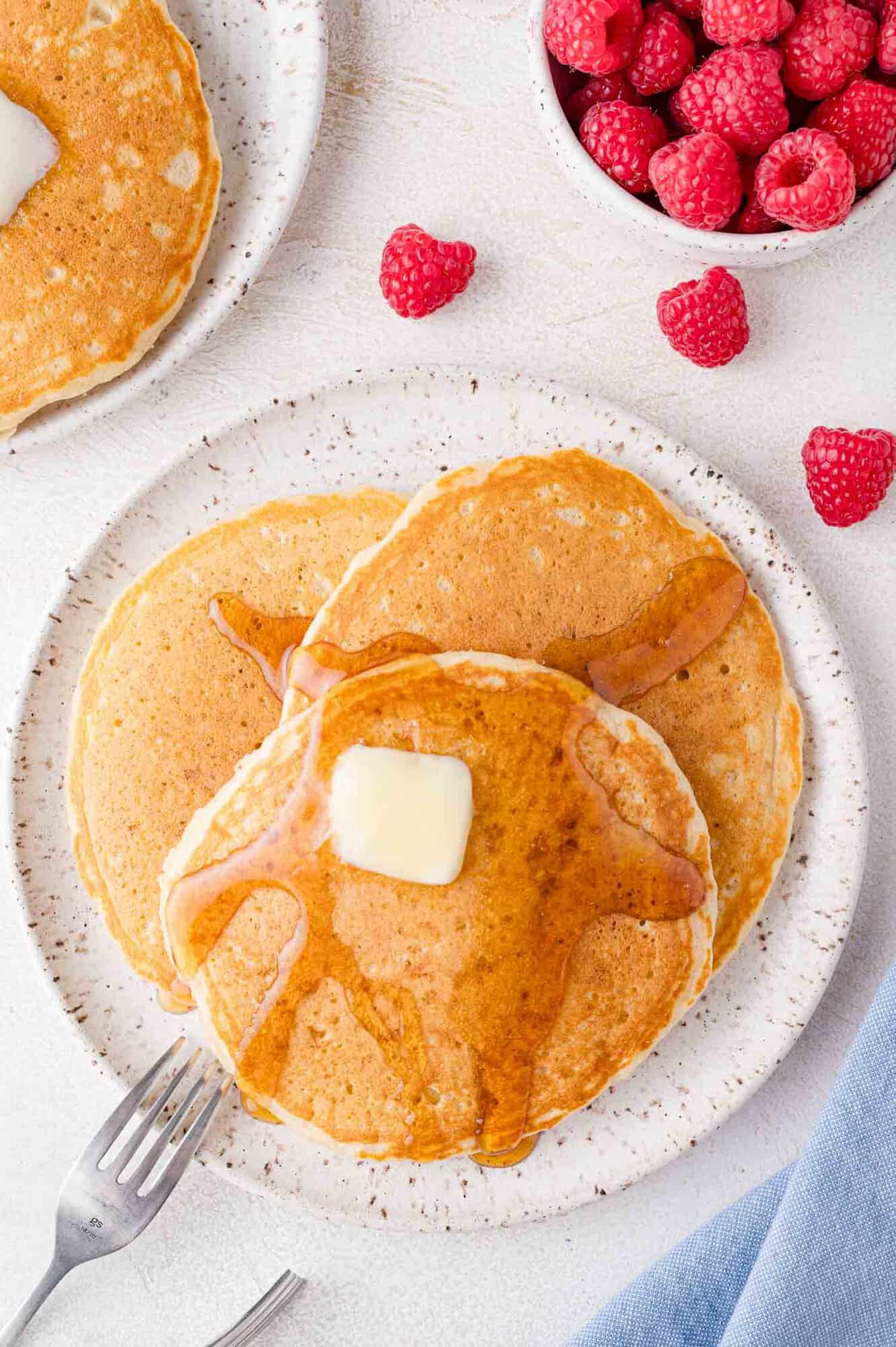 Pancakes, plated, with butter and syrup. Raspberries in the corner of the photo.