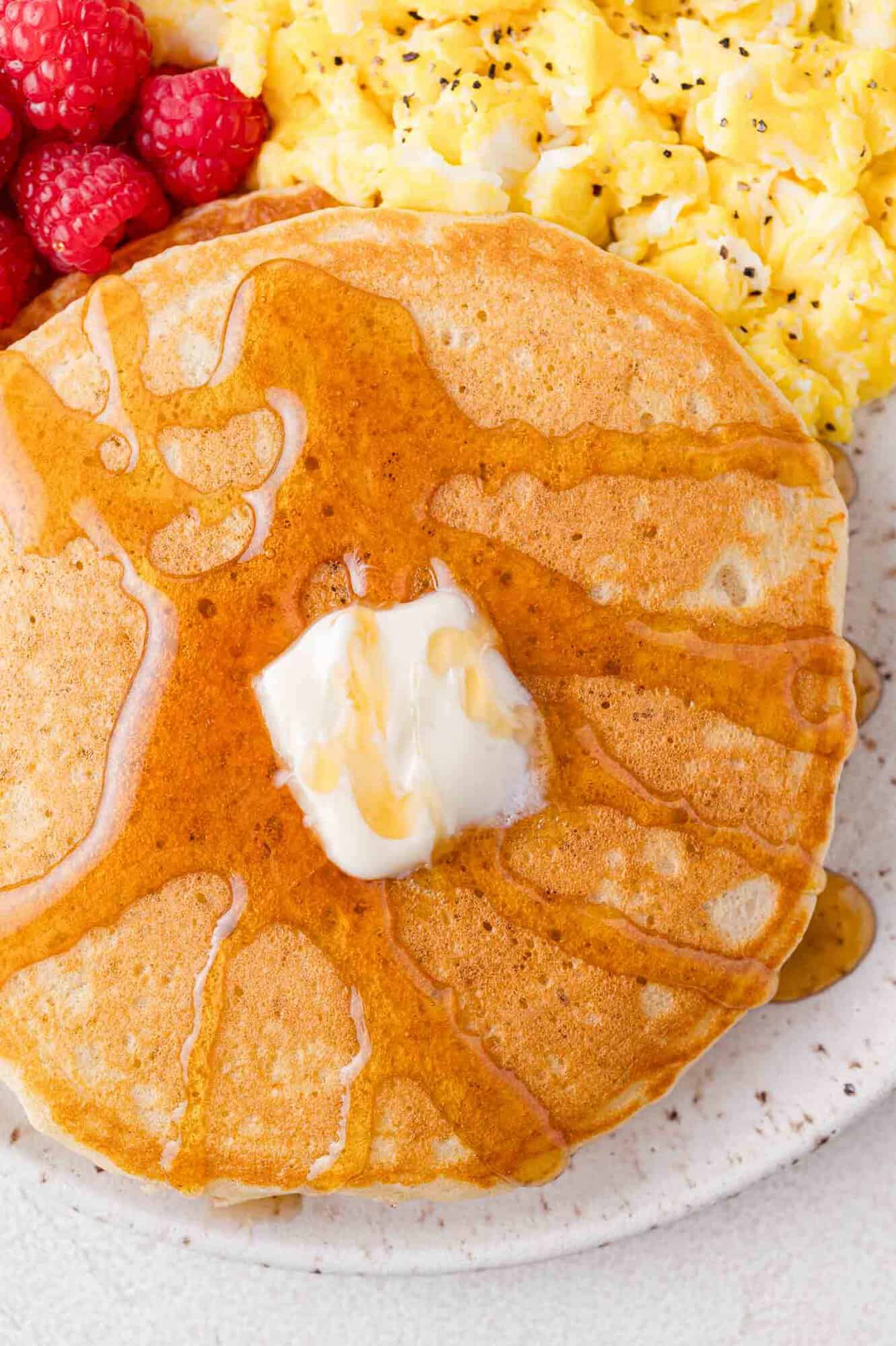 Close up of butter and syrup on pancakes, on a plate with eggs and raspberries.