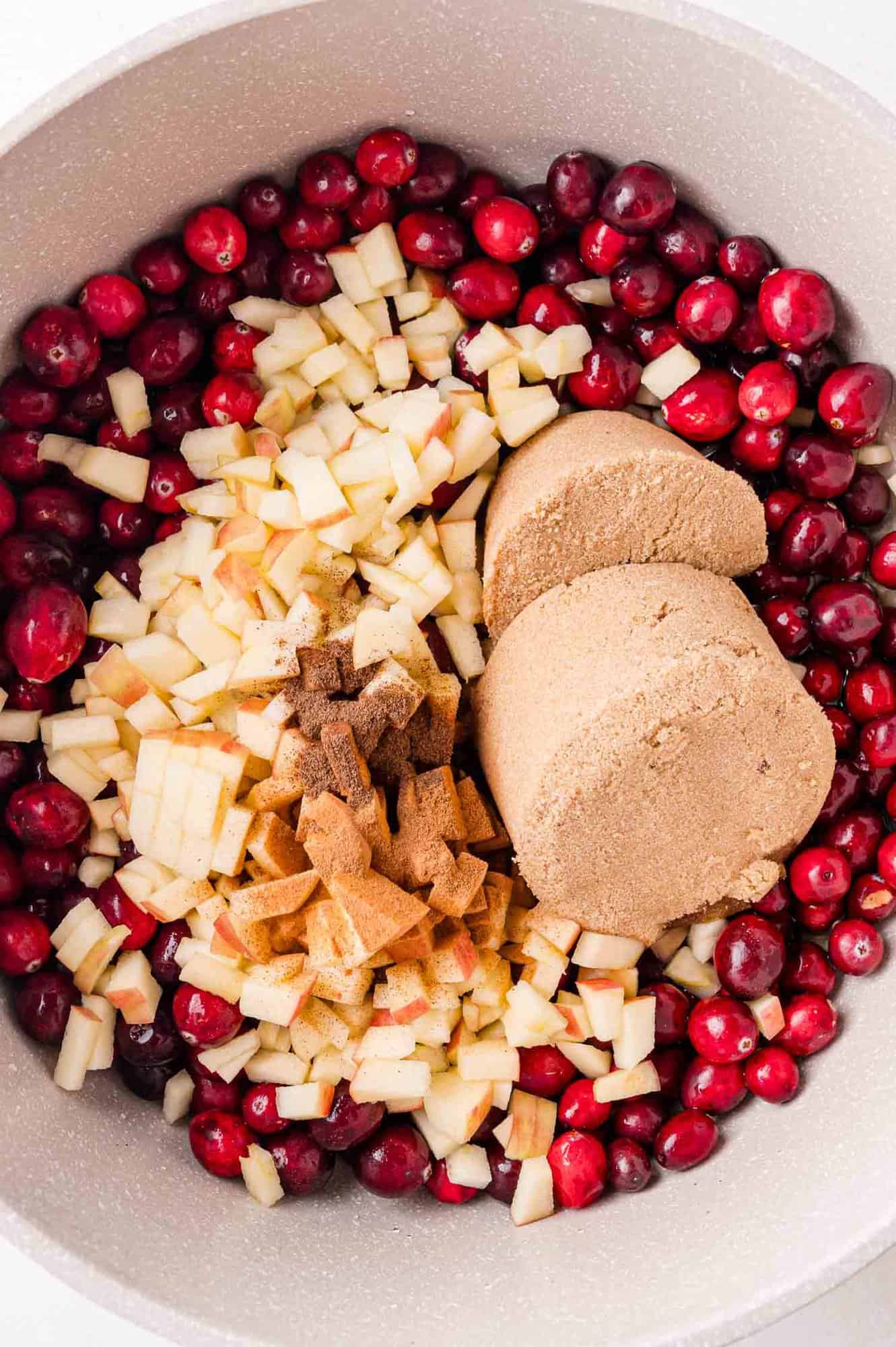 A large mixing bowl filled with fresh cranberries, diced apples, brown sugar, cinnamon and nutmeg, ready to be mixed for a recipe.