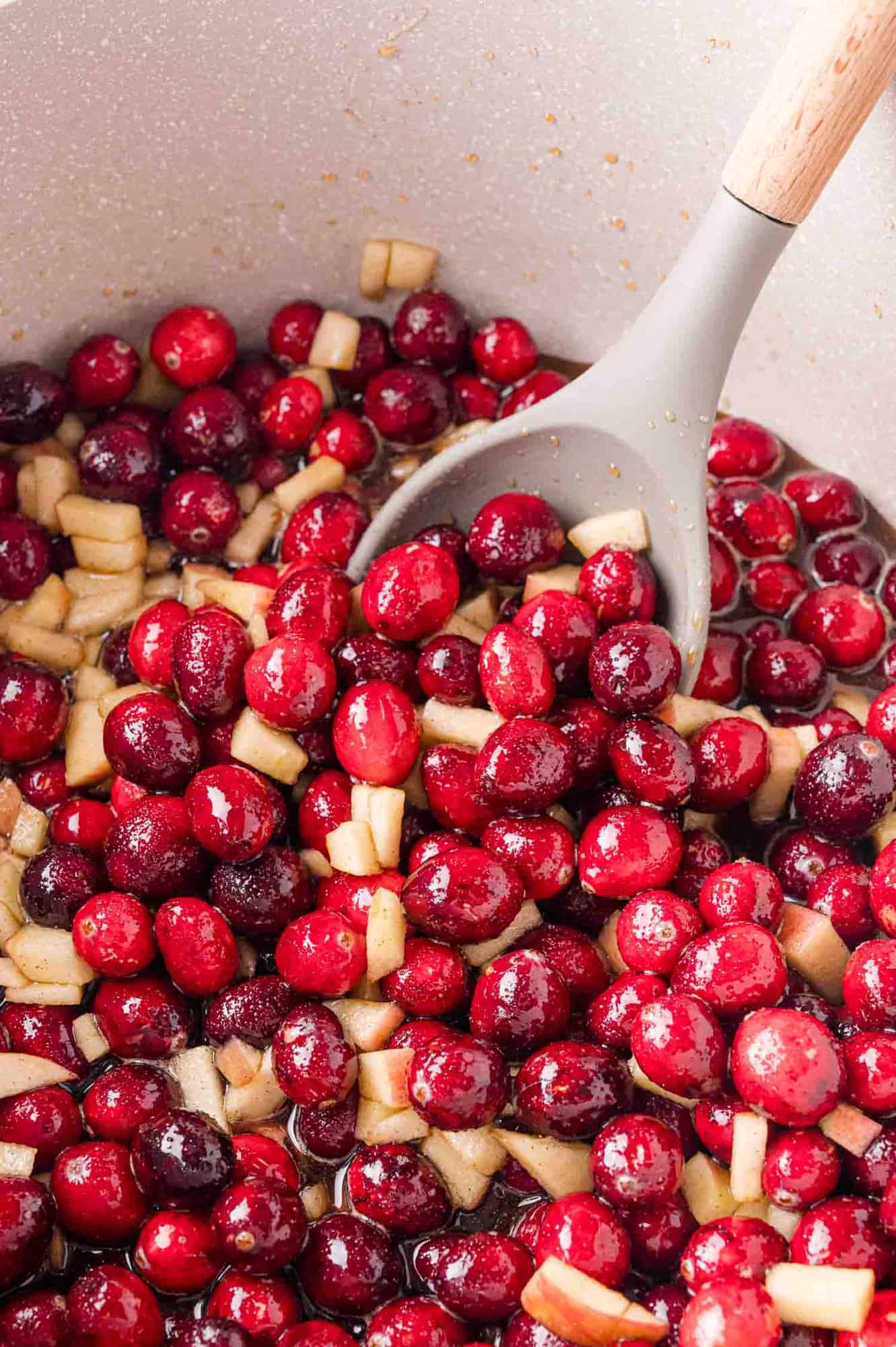 A pot filled with red cranberries and apple pieces, being stirred with a grey spoon. 