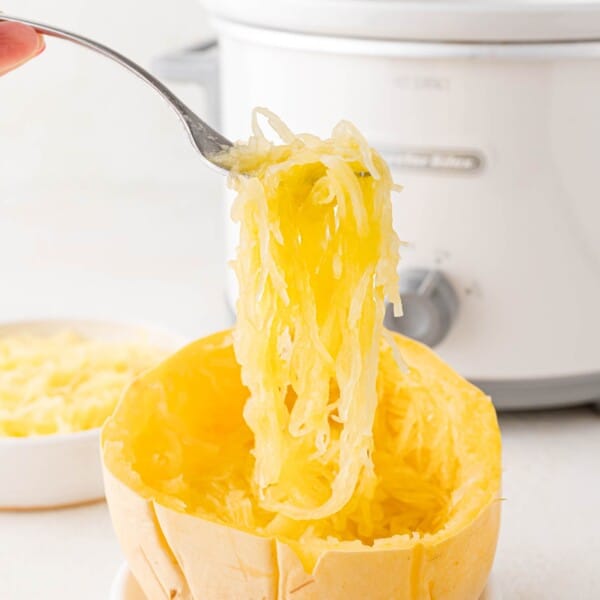 Spaghetti squash strands being lifted out of the squash half with a fork. Crockpot in background.