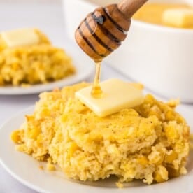 Corn spoon bread scooped onto a plate, with butter on top, being drizzled with honey.