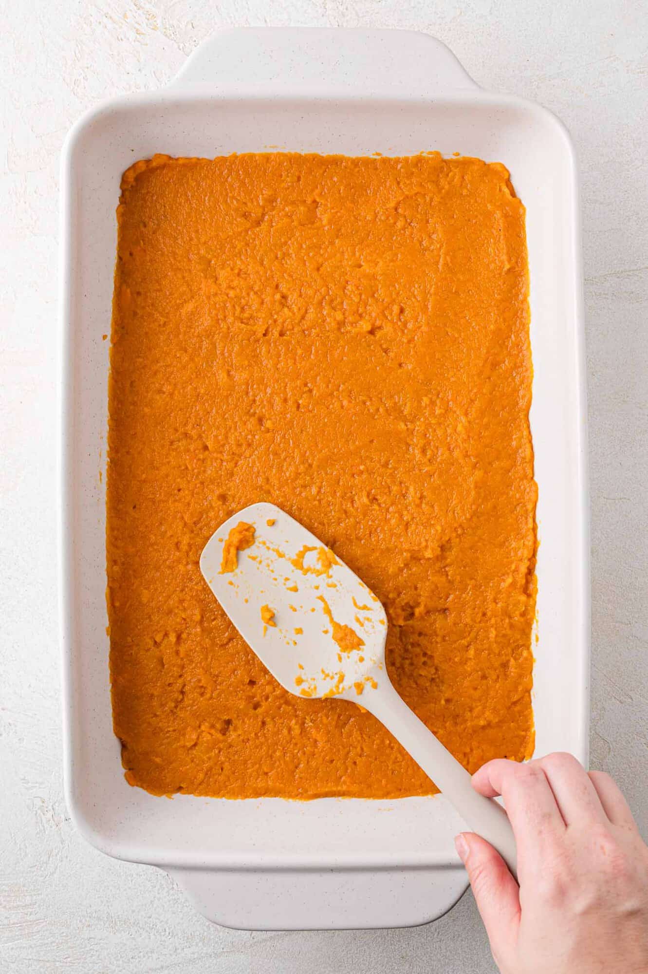 Sweet potato casserole being spread into white casserole dish.