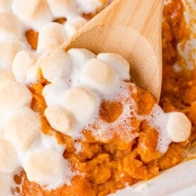 Sweet potato casserole, topped with marshmallows, being scooped with a wooden spoon.