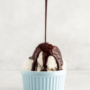 Chocolate syrup being poured onto vanilla ice cream in a light blue bowl against a plain background.