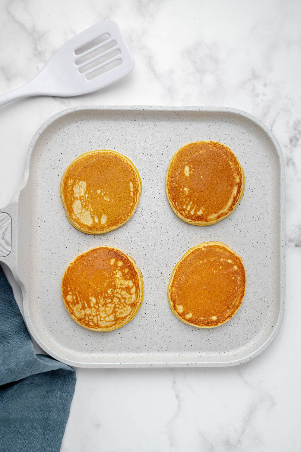 Four golden-brown pancakes on a white griddle. A gray spatula and blue napkin are nearby on a marble surface.