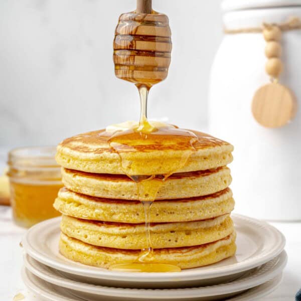 A stack of pancakes on a plate with butter on top, being drizzled with honey. A jar of honey and a white container are in the background.