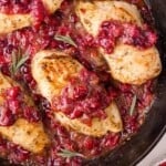 Chicken breasts in a skillet topped with cranberry sauce, garnished with rosemary sprigs.