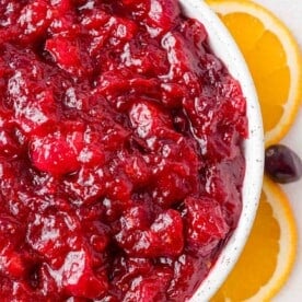 A close-up of a bowl filled with cranberry sauce, surrounded by slices of orange.