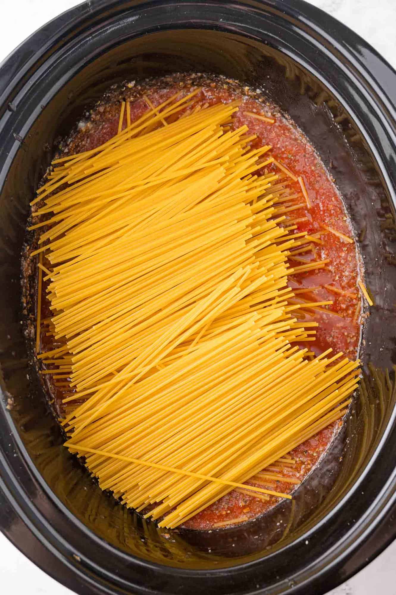 Uncooked spaghetti placed in a slow cooker filled with tomato sauce.