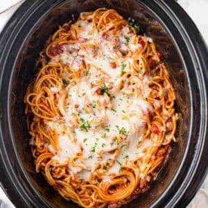 Spaghetti with tomato sauce and melted cheese in a black slow cooker, topped with chopped parsley.