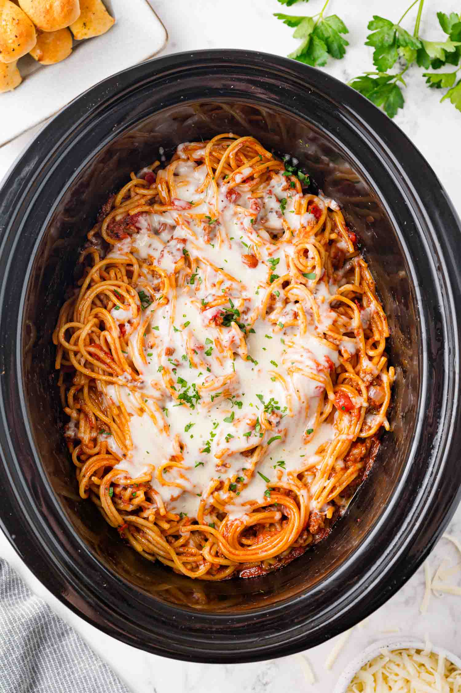 Spaghetti with tomato sauce and melted cheese in a black slow cooker, topped with chopped parsley.