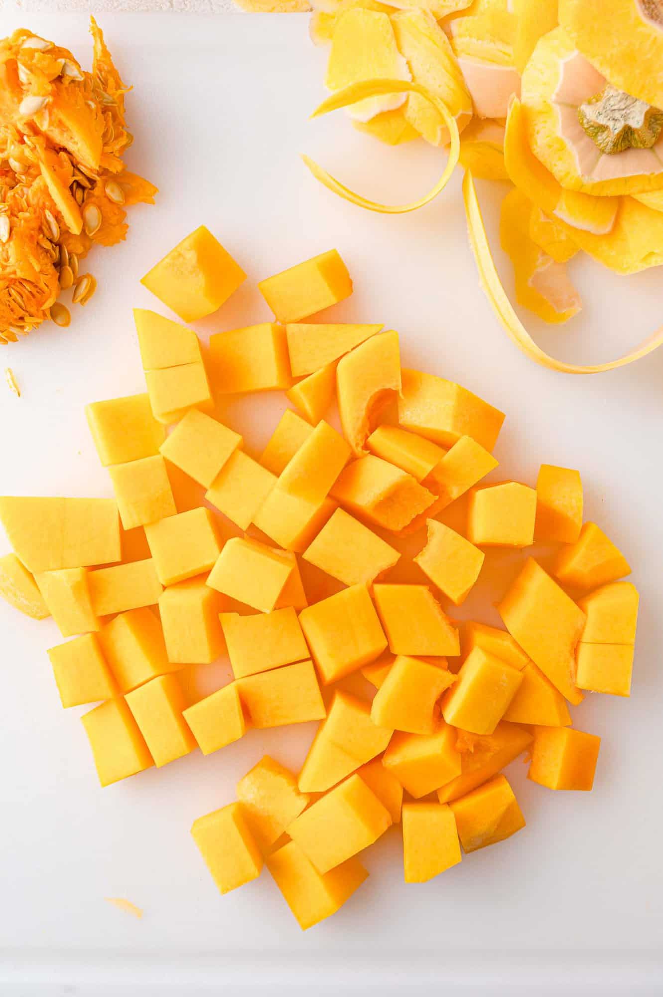 Cubed butternut squash with peelings and seeds on a white cutting board.