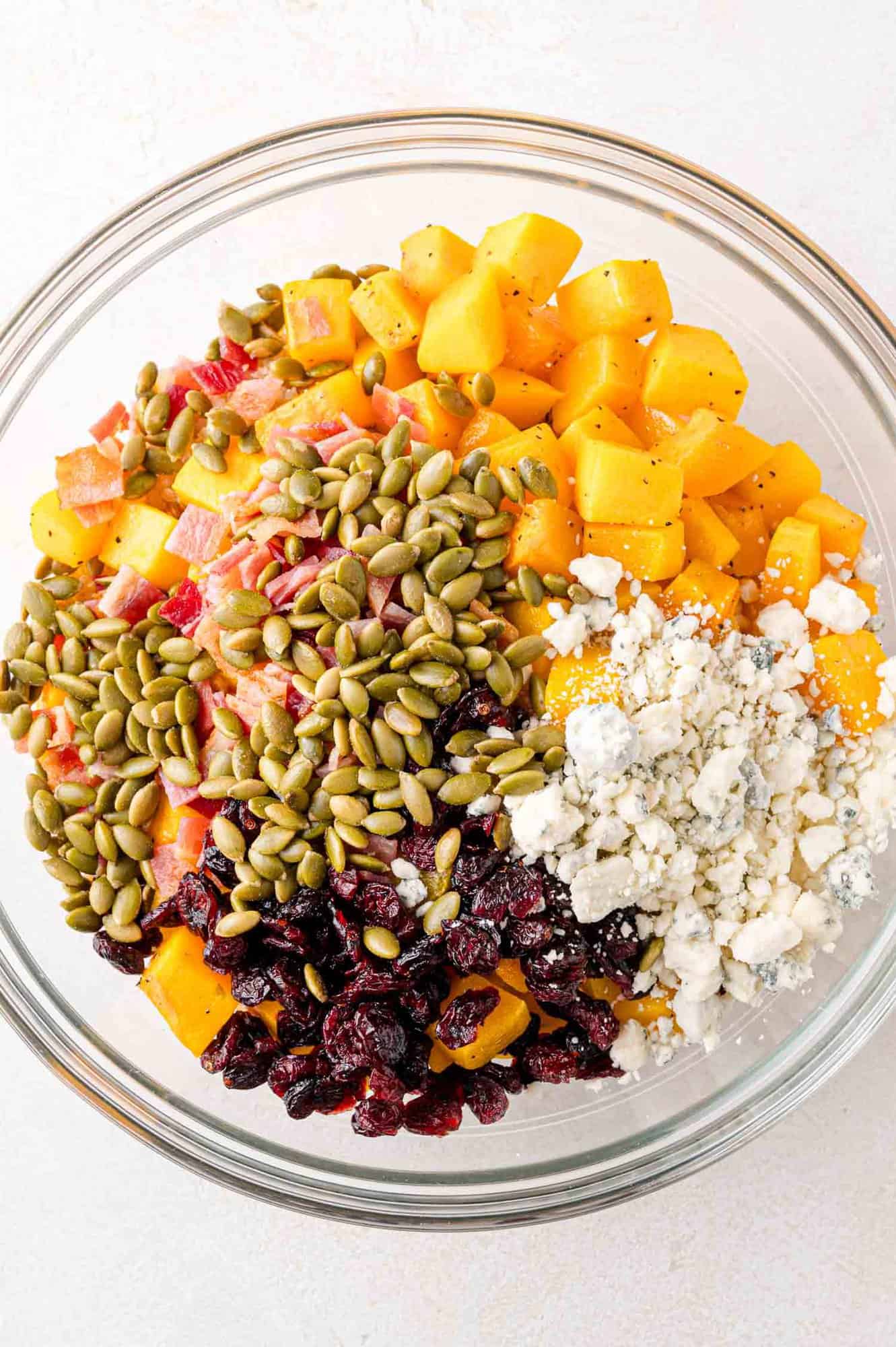 A bowl contains diced butternut squash, pepitas, cranberries, and crumbled cheese.