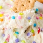 Close-up of a creamy, white dip with colorful sprinkles and a graham cracker partially inserted on top.