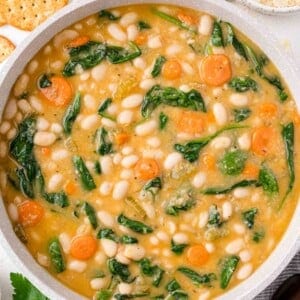 A pot of soup with white beans, carrots, and spinach. Crackers, parsley, and two spoons are nearby.