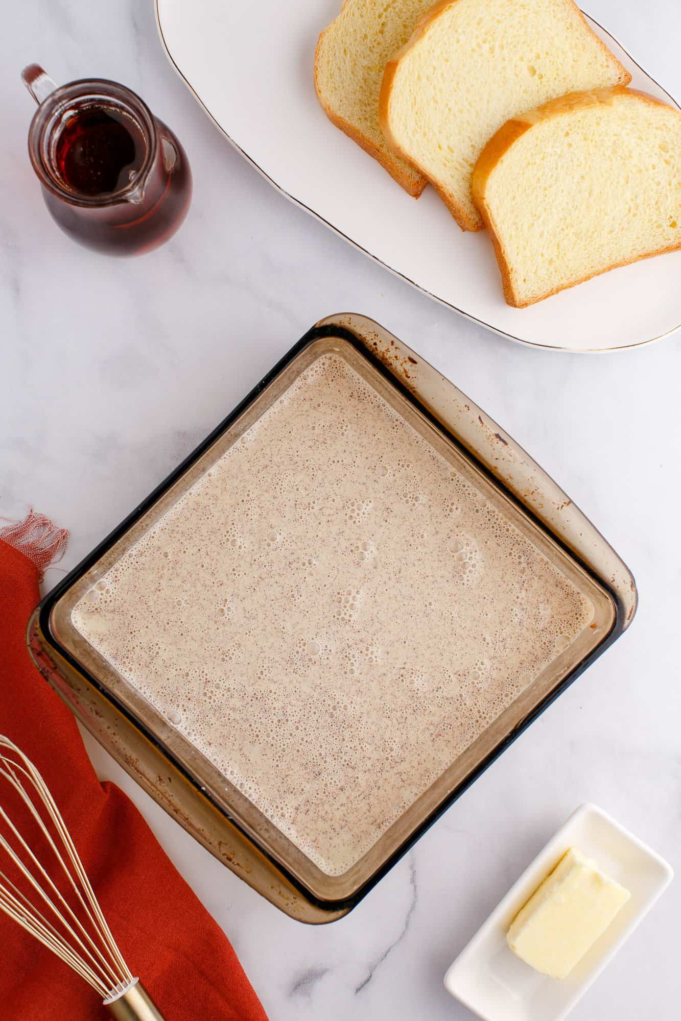 A glass baking dish with a light brown mixture, a plate with sliced bread, a small pitcher of liquid, a stick of butter, a whisk, and an orange cloth on a marble surface.