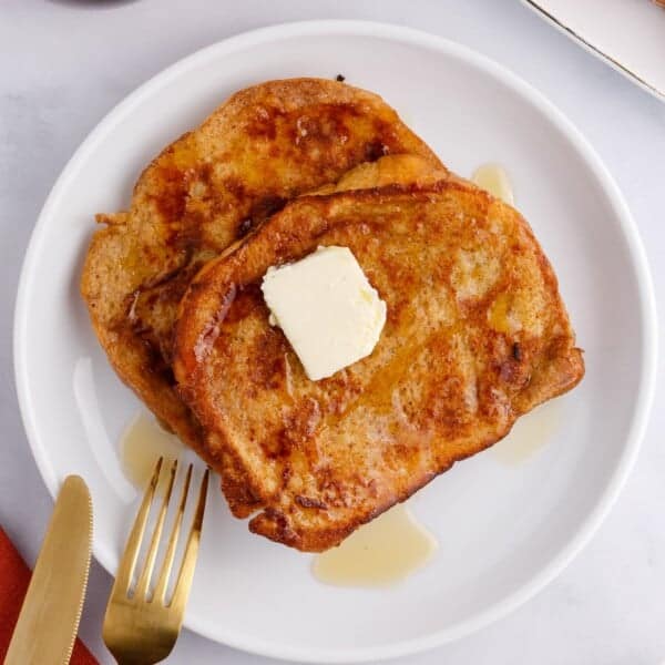 Two slices of brioche French toast with a pat of butter and syrup on a white plate, next to a gold fork.