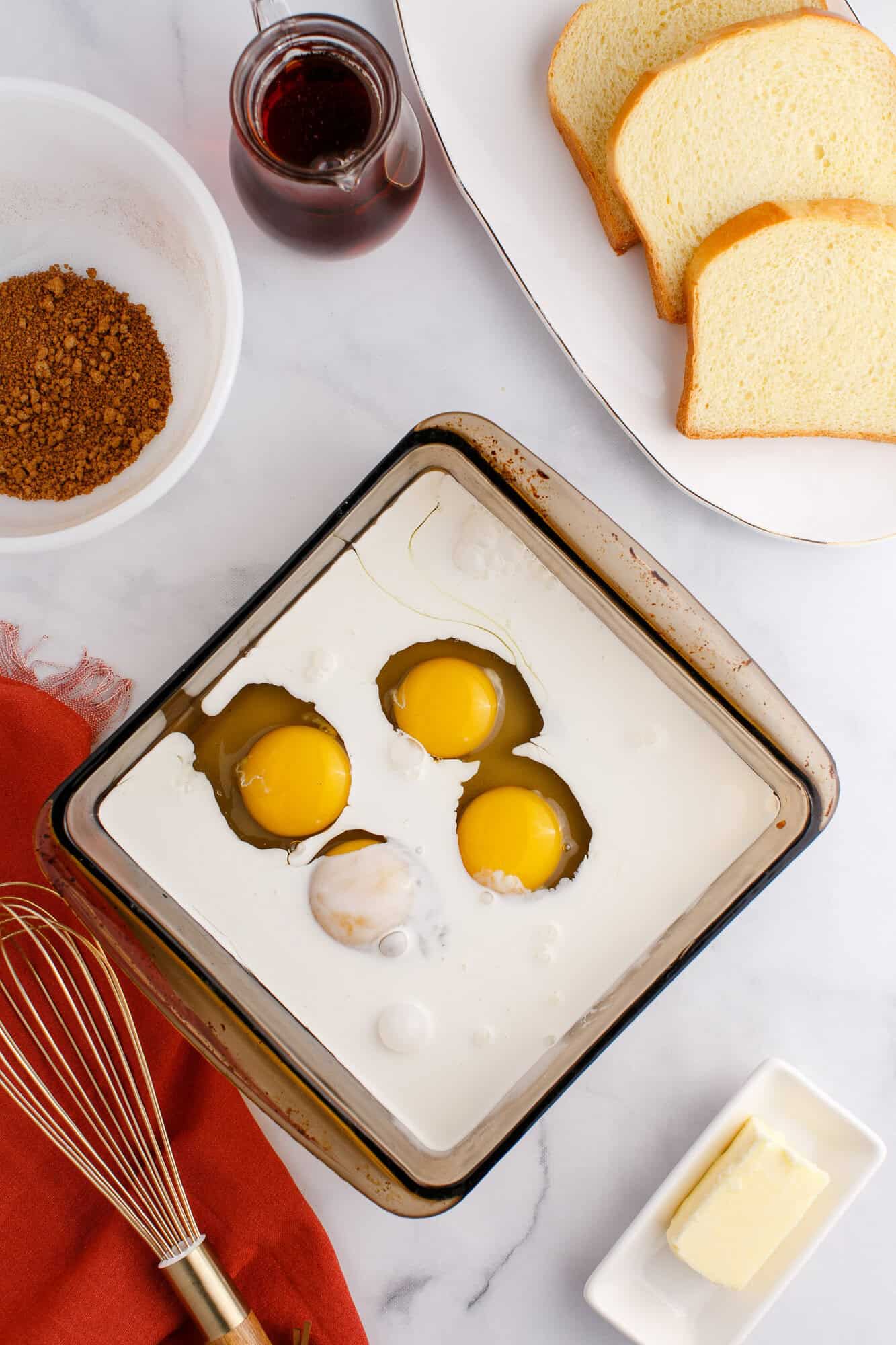 A baking dish with cream and three cracked eggs, slices of bread on a plate, and a small jar of syrup. A bowl of brown sugar and butter are nearby on a marble countertop.
