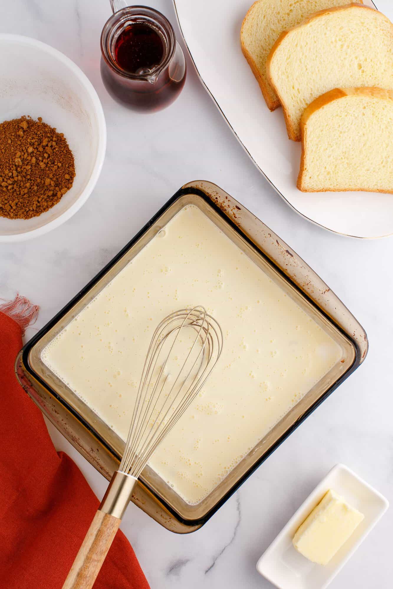 Ingredients for making French toast, including bread slices, a whisk in a bowl of batter, a bowl of spices, a dish of butter, and a jar of syrup on a marble surface.