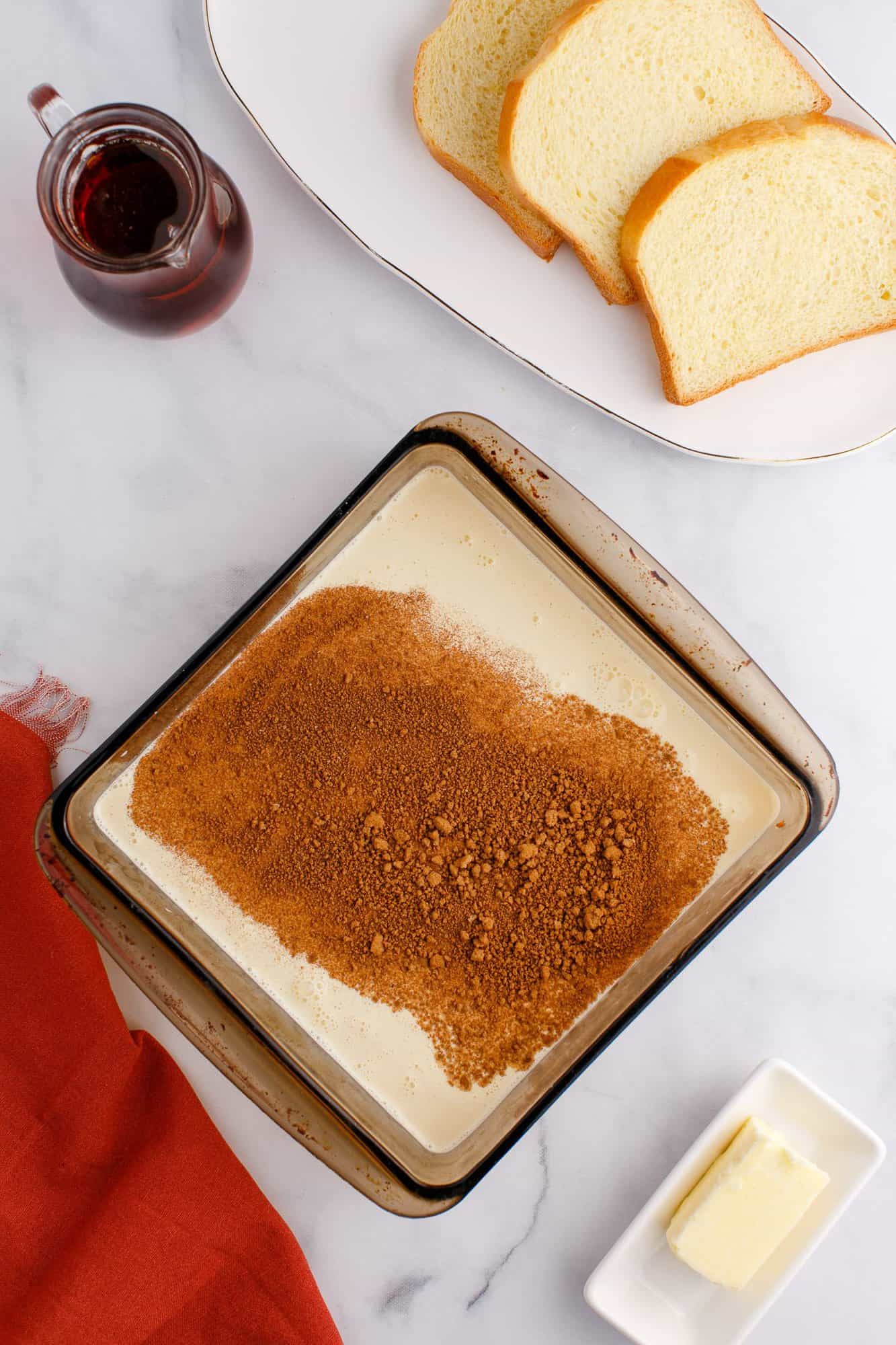 A baking dish with layered cream and brown sugar, alongside two slices of bread, a small pitcher of syrup, and a dish with a butter stick on a marble surface.