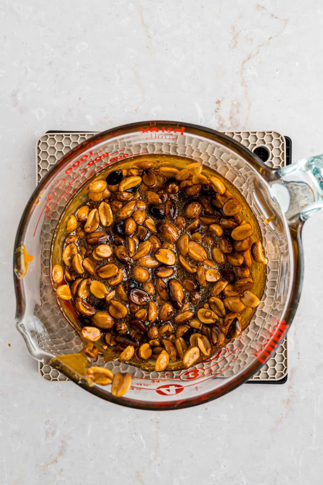 Top view of peanuts and burnt candy in a glass measuring cup on a light surface.