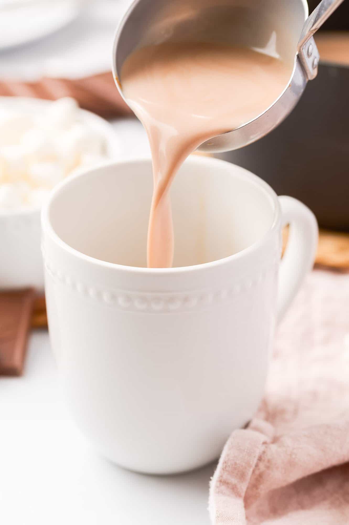 Pouring creamy hot chocolate from a ladle into a white mug, with marshmallows and a pink napkin nearby.