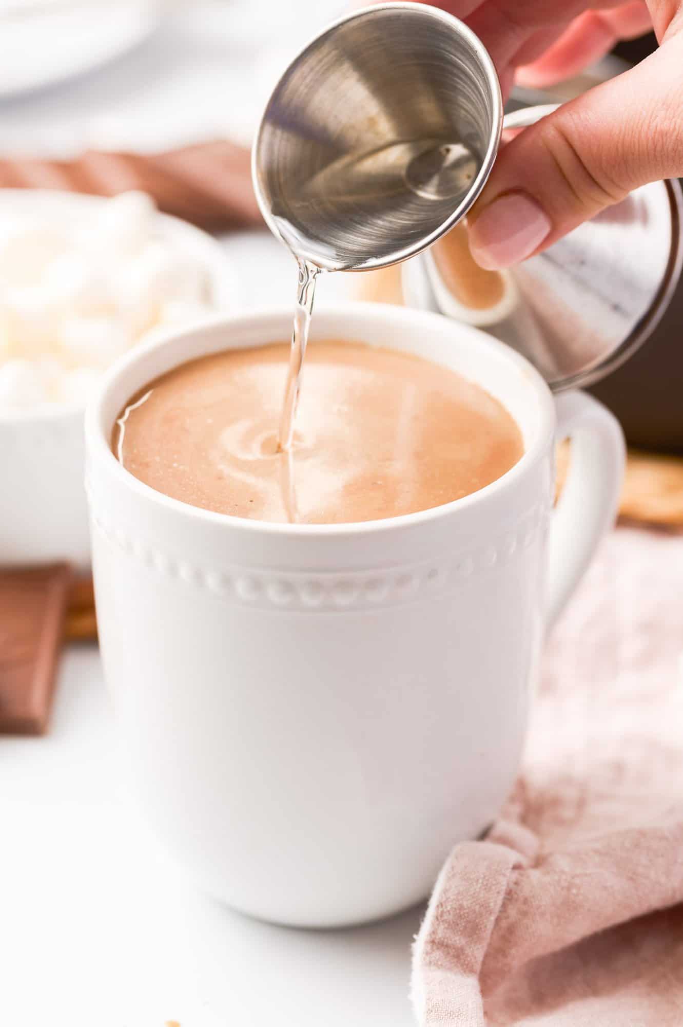 Pouring liquid from a small metal cup into a mug of creamy coffee on a table with a light pink cloth.
