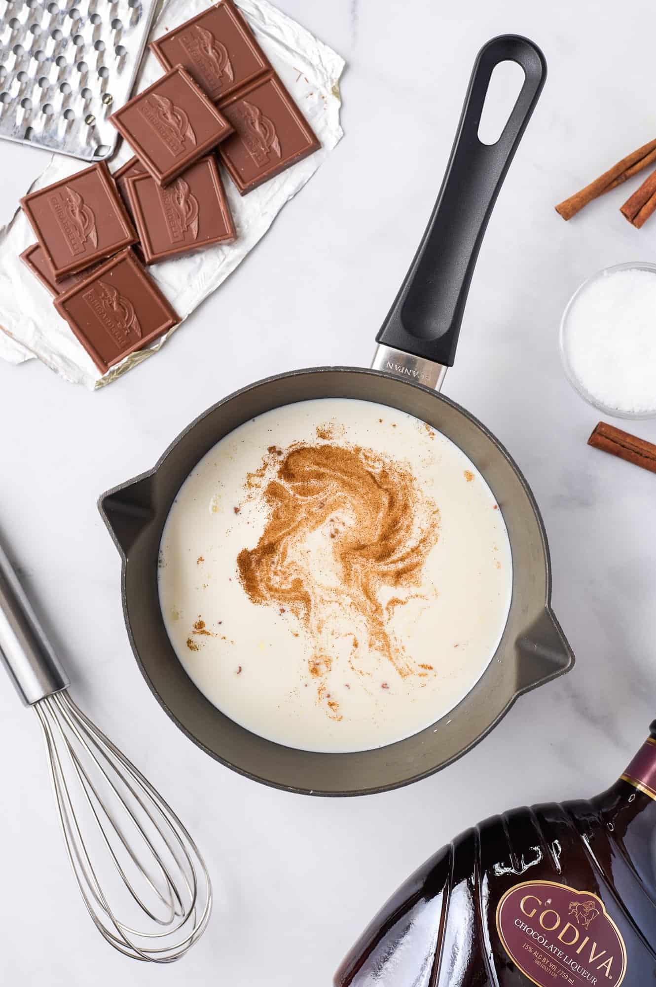 A saucepan with milk and cinnamon on a stovetop, surrounded by chocolate pieces, sugar, a whisk, and a bottle of chocolate liqueur.