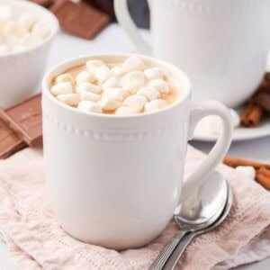 A white mug filled with hot chocolate topped with mini marshmallows, placed on a pink cloth with a spoon beside it.
