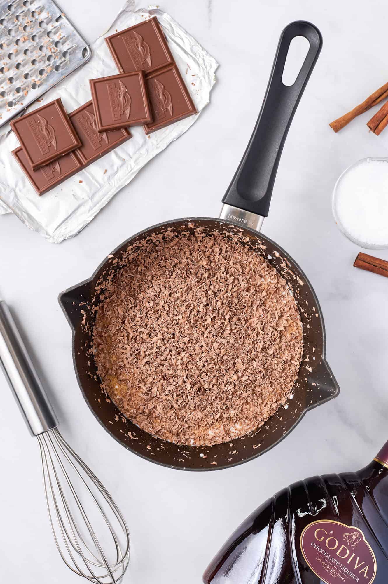 A saucepan with grated chocolate on a marble surface, surrounded by chocolate bars, a whisk, Godiva liqueur bottle, a bowl of sugar, and cinnamon sticks.