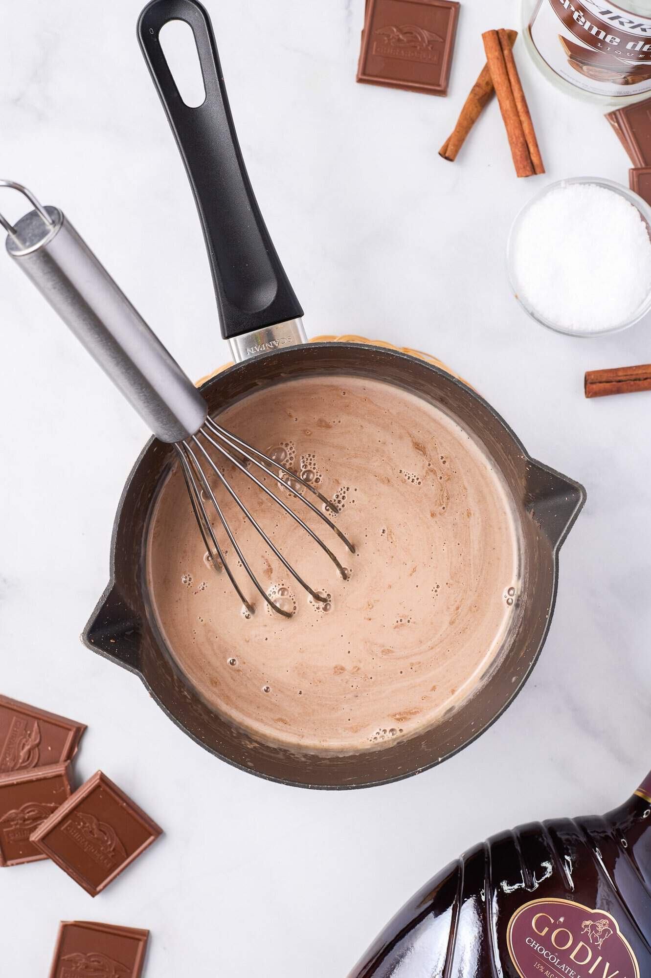 A saucepan with chocolate milk being whisked, surrounded by chocolate pieces, cinnamon sticks, and a sugar bowl on a marble surface.
