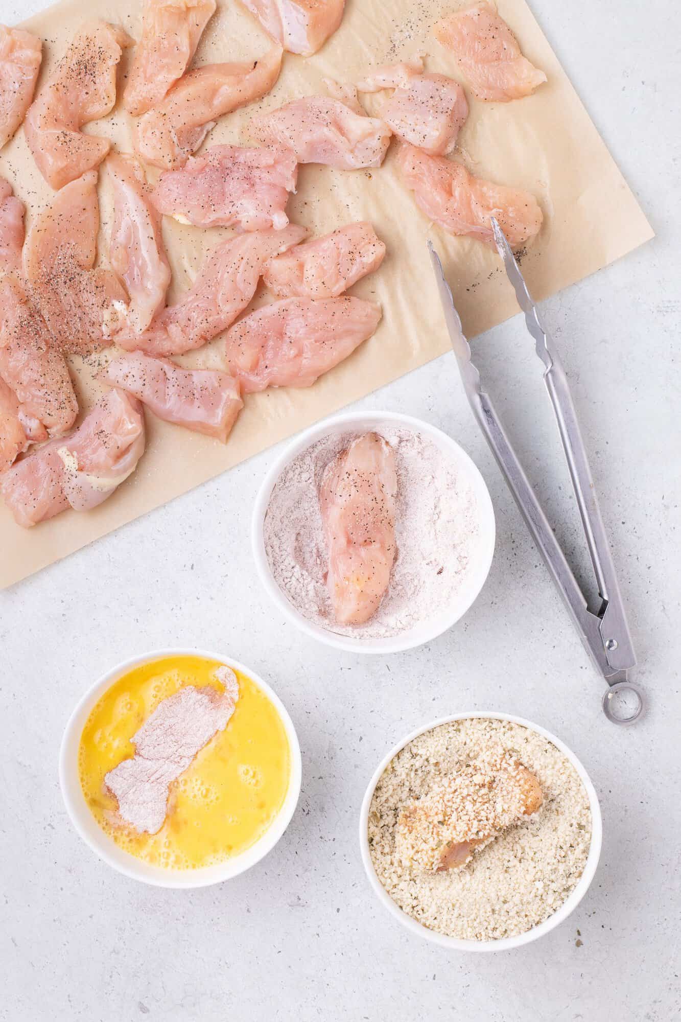 Raw chicken strips are on parchment paper. Nearby are bowls with flour, beaten eggs, and breadcrumbs for breading. Metal tongs lie beside them on a white surface.