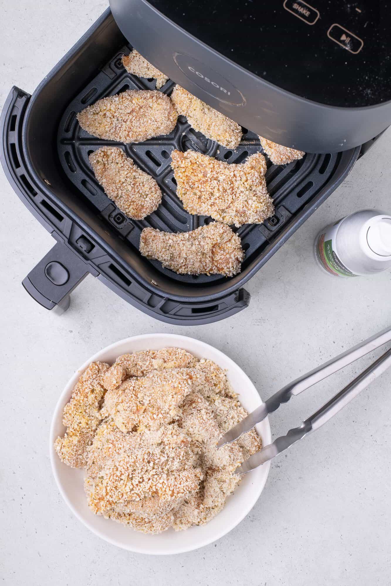 Air fryer with breaded chicken pieces inside. A bowl with more breaded chicken and metal tongs is beside it.
