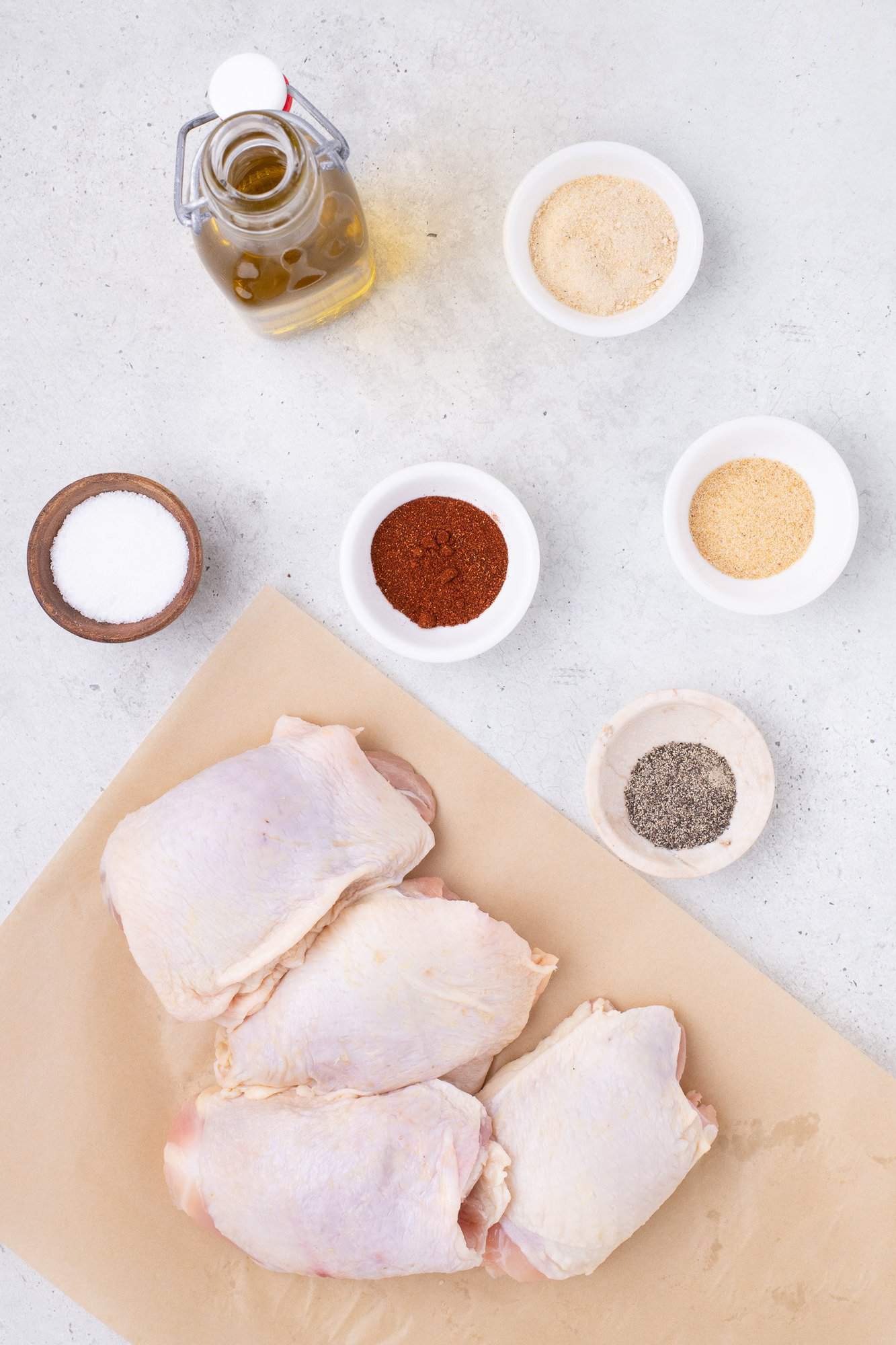 Raw chicken thighs on parchment paper with olive oil, spices, and seasonings in small bowls on a light background.