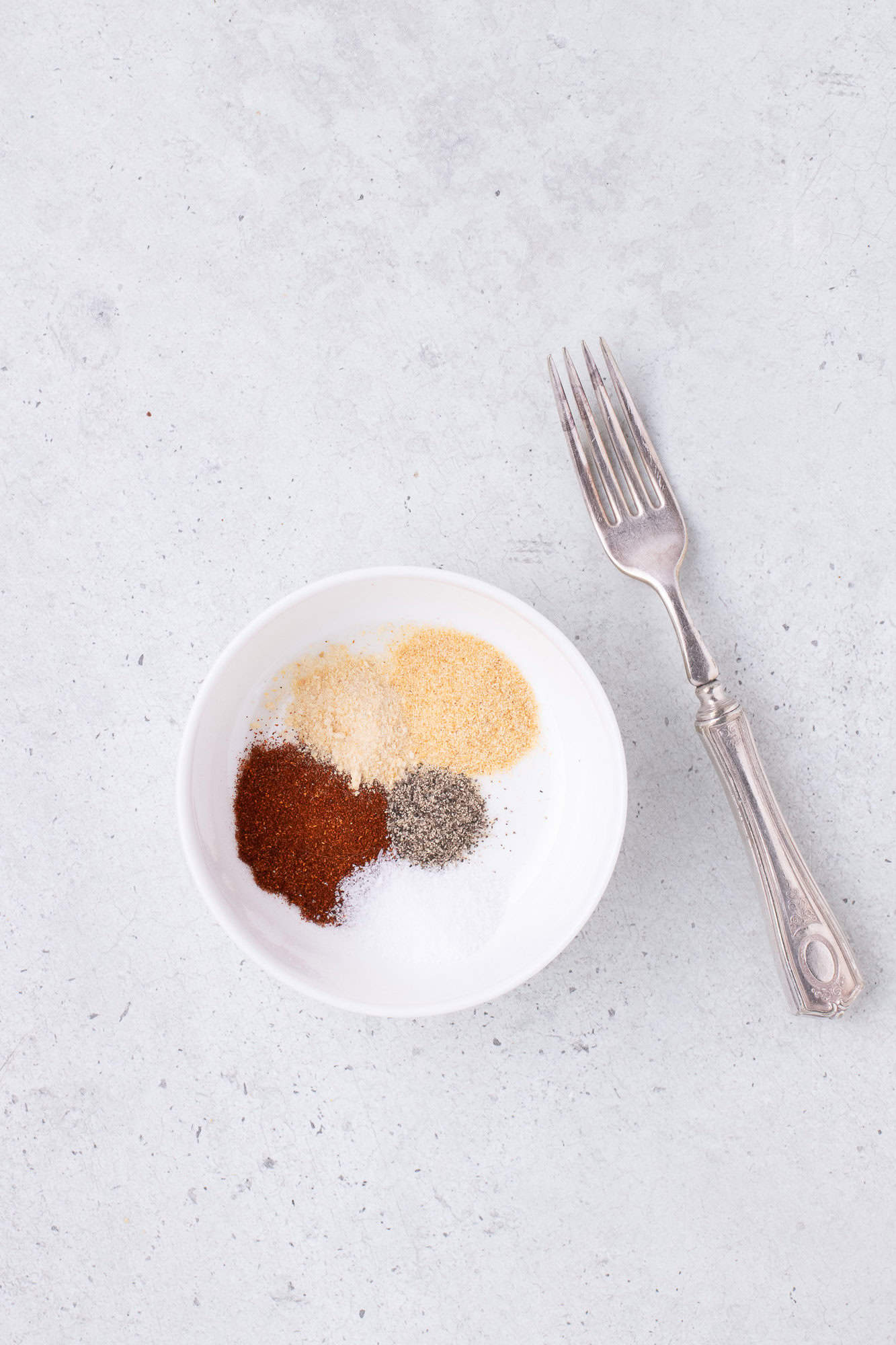 A small white bowl with assorted spices and a silver fork on a light gray surface.