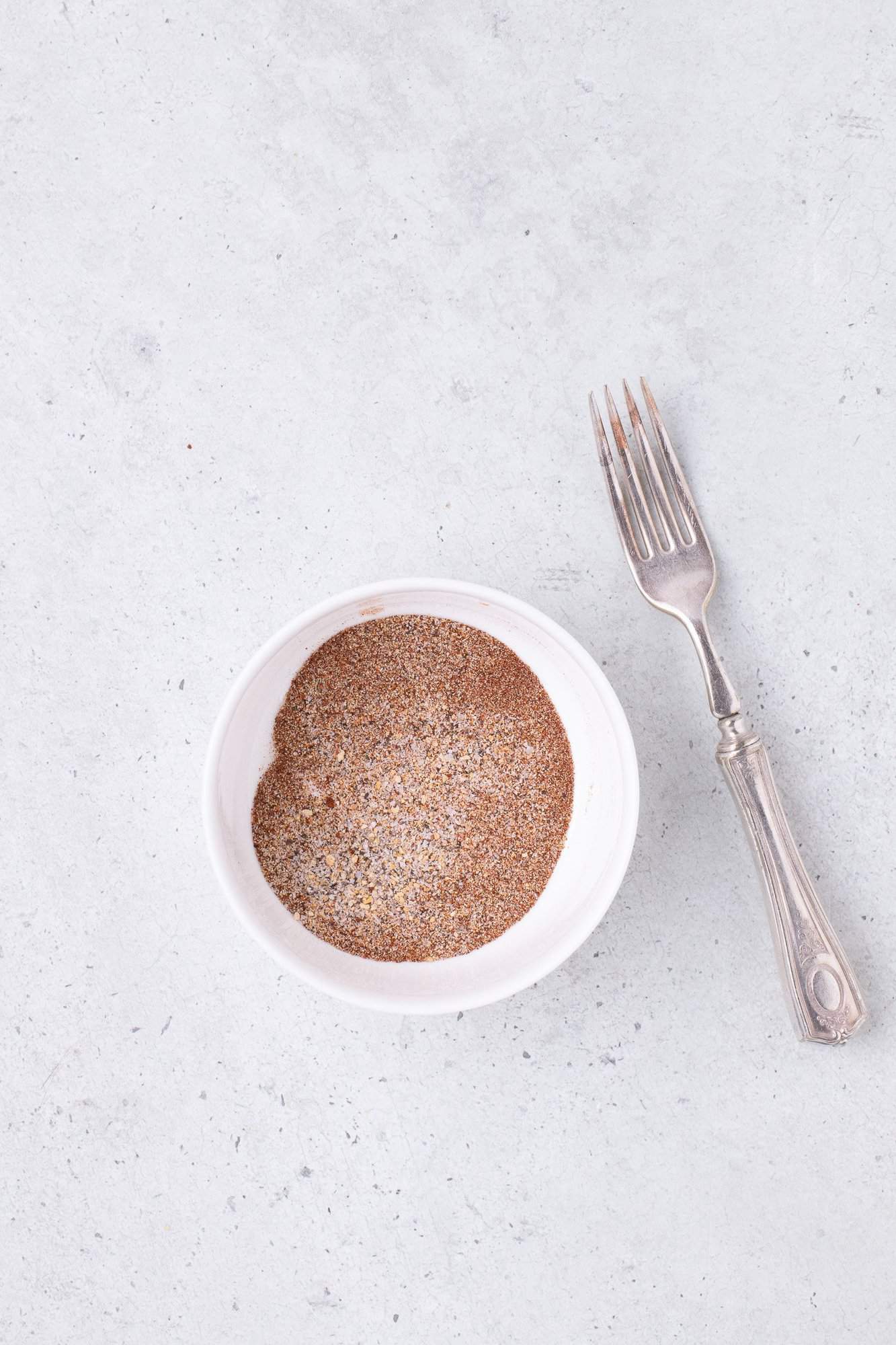 A white bowl filled with ground cinnamon and sugar mixture next to a silver fork on a light gray surface.