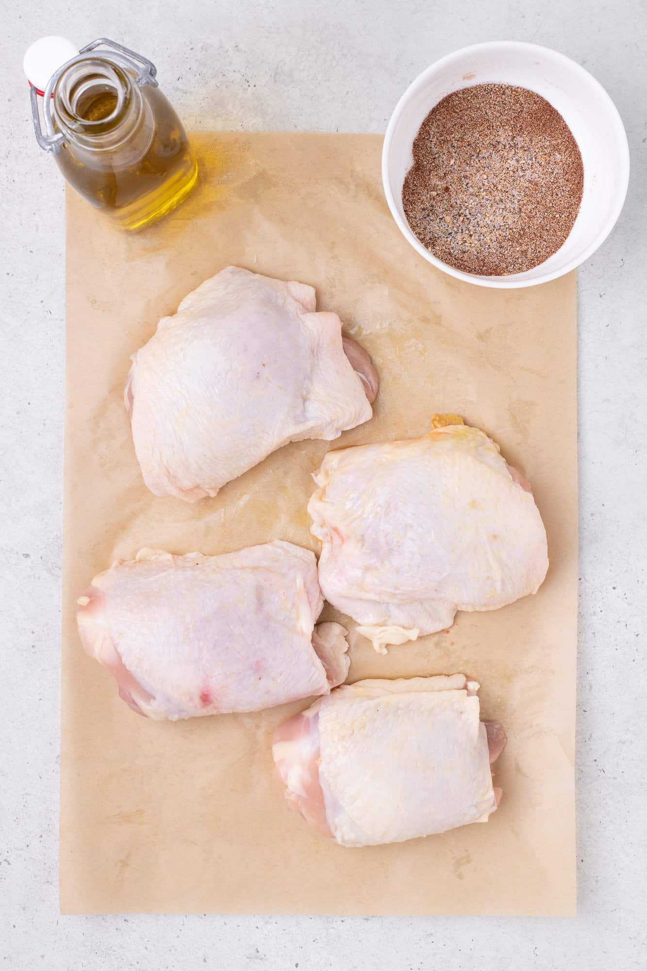 Four raw chicken thighs on parchment paper with a bowl of seasoning and a bottle of oil.