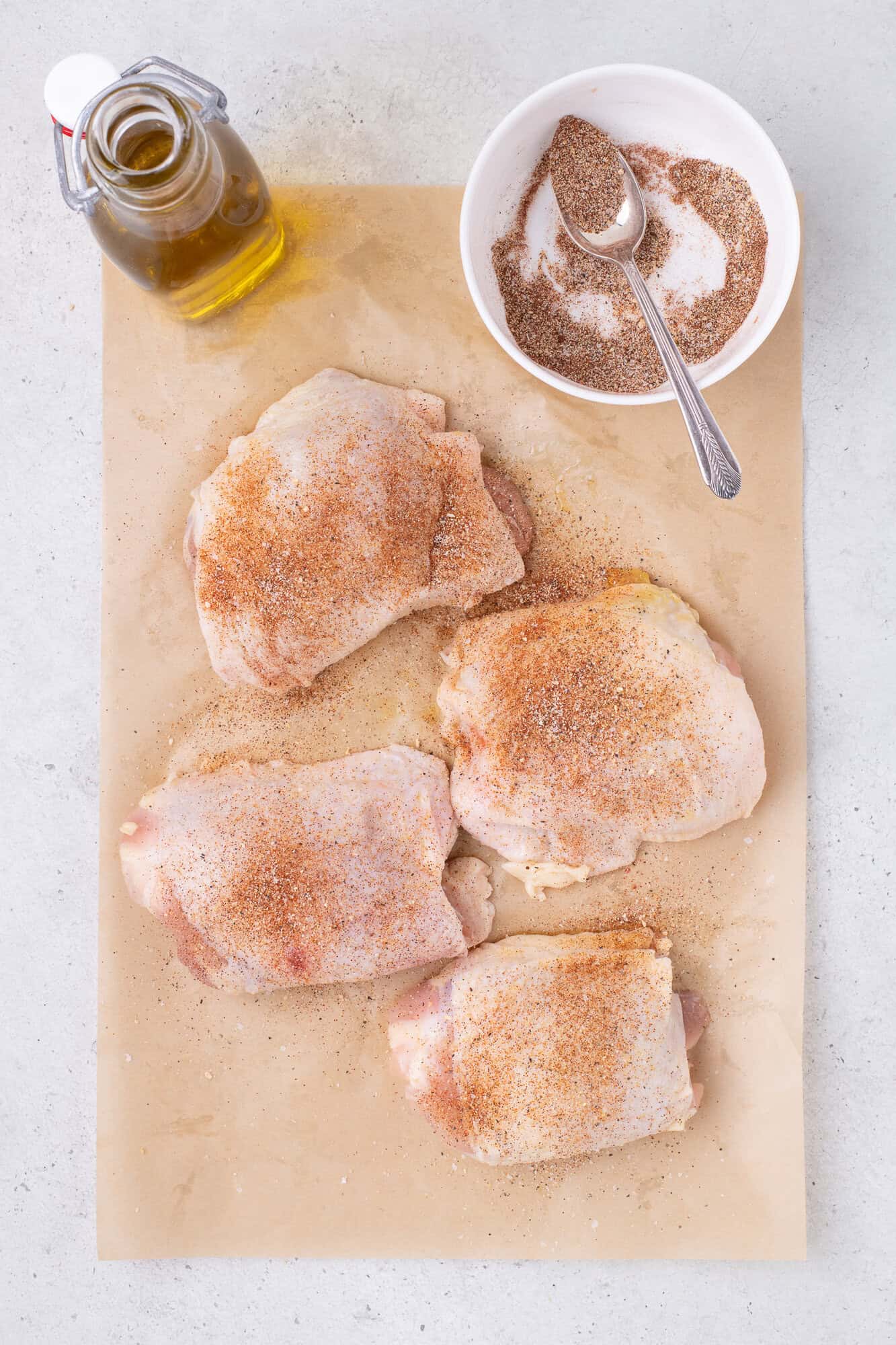 Raw chicken thighs seasoned with spices on parchment paper, next to a small bowl of spice mix with a spoon and a bottle of oil.